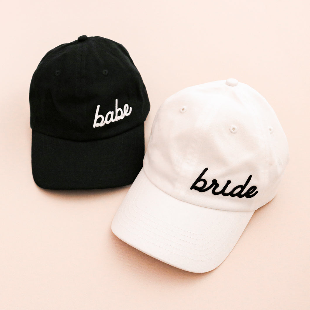 Black baseball cap with the word babe in white script. White cap with the word bride in black script. 