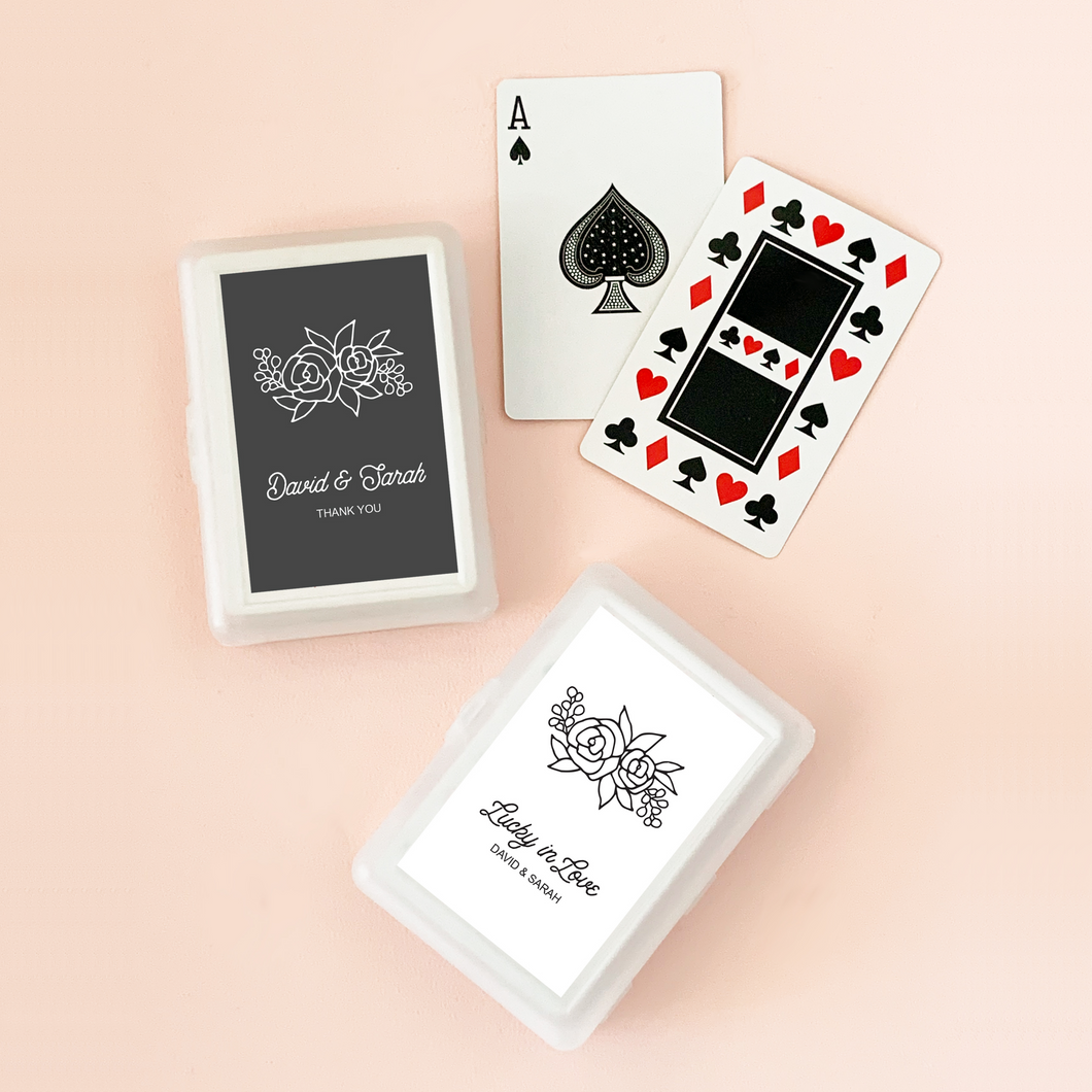 Two deck of cards in plastic cases, each case with a customized label of two lines of text and a floral design. One label shown in black and the other in white.