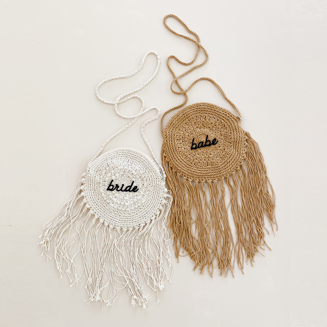 Two circle crochet bags with hanging fringe. One in ivory with the word bride in black script in the center. The other in natural tan with the word babe in black script in the center.