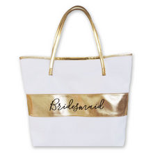 Load image into Gallery viewer, White tote bag with a gold handle and horizontal gold strip on the middle of the body of the bag. The word &quot;bridesmaid&quot; in black script is on the gold strip.
